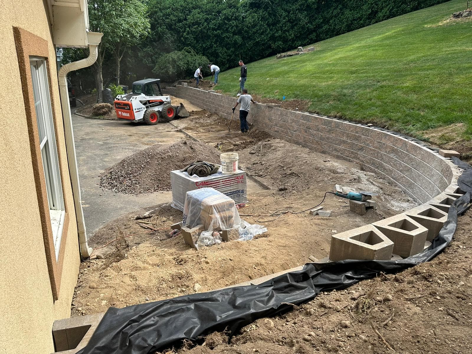 Workers building a curved stone retaining wall on a sloped lawn with a Bobcat.