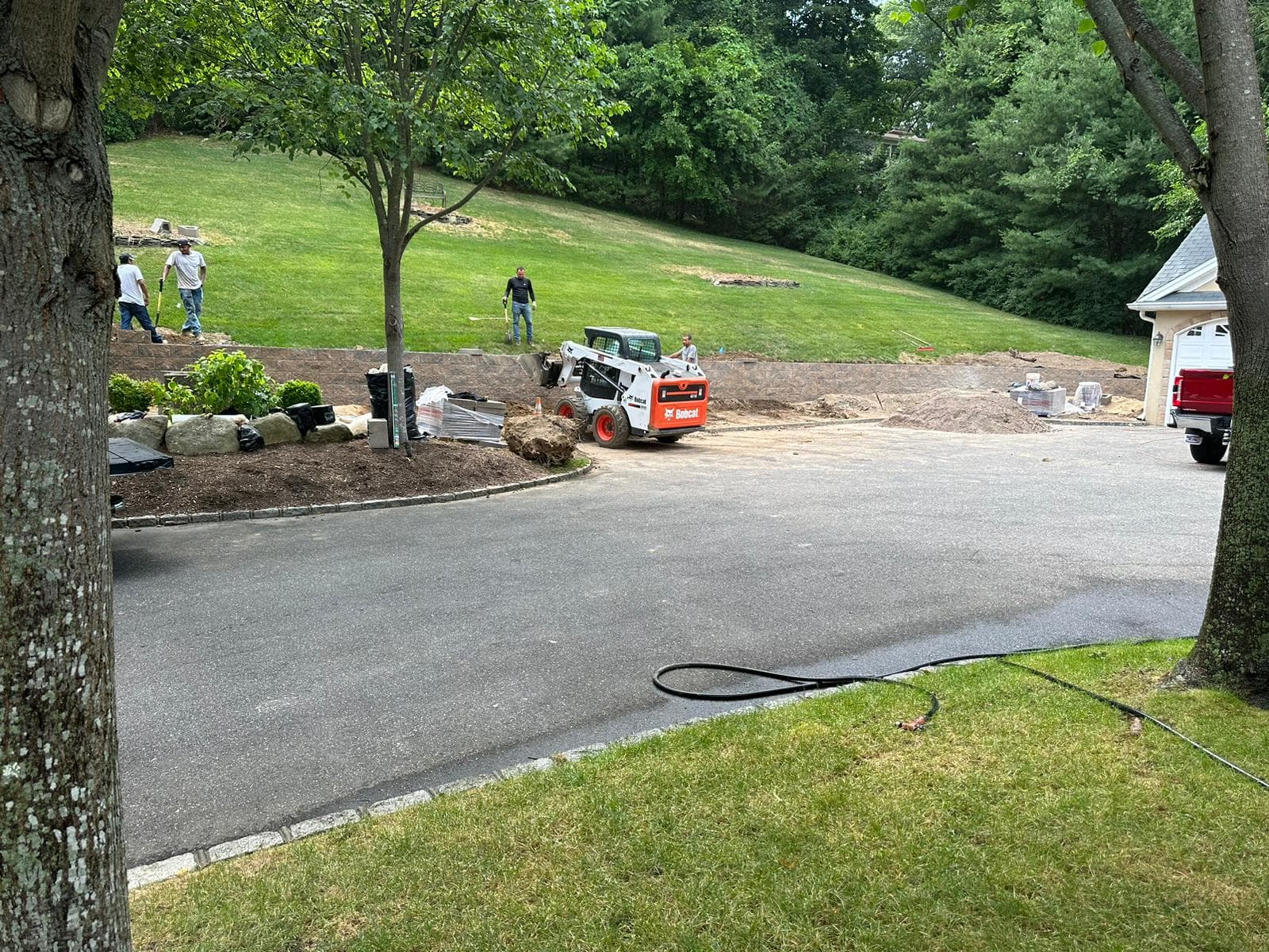A Bobcat loader and workers build a stone retaining wall along a residential driveway.