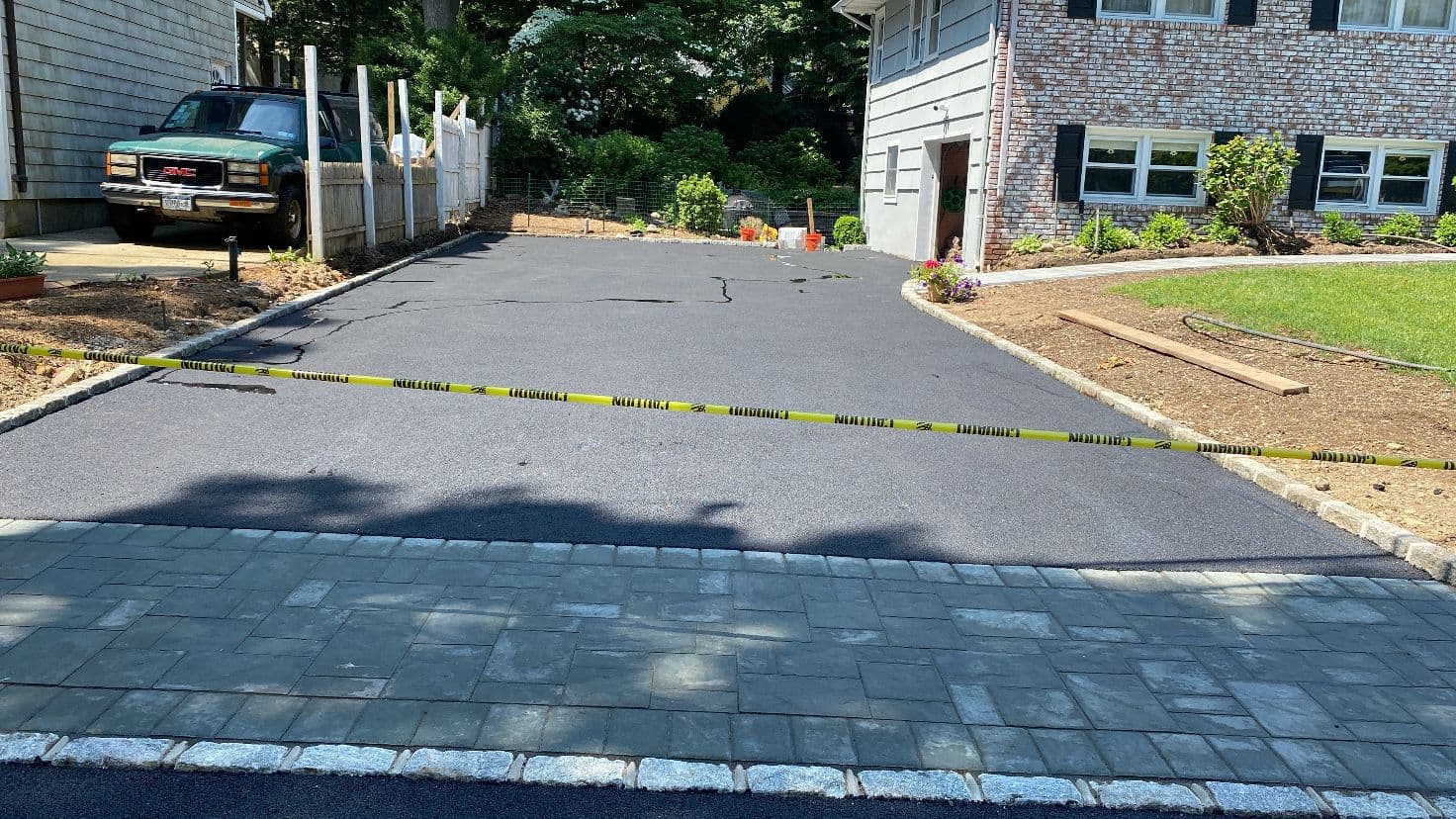 Newly paved asphalt driveway with yellow caution tape and a decorative stone paver apron.
