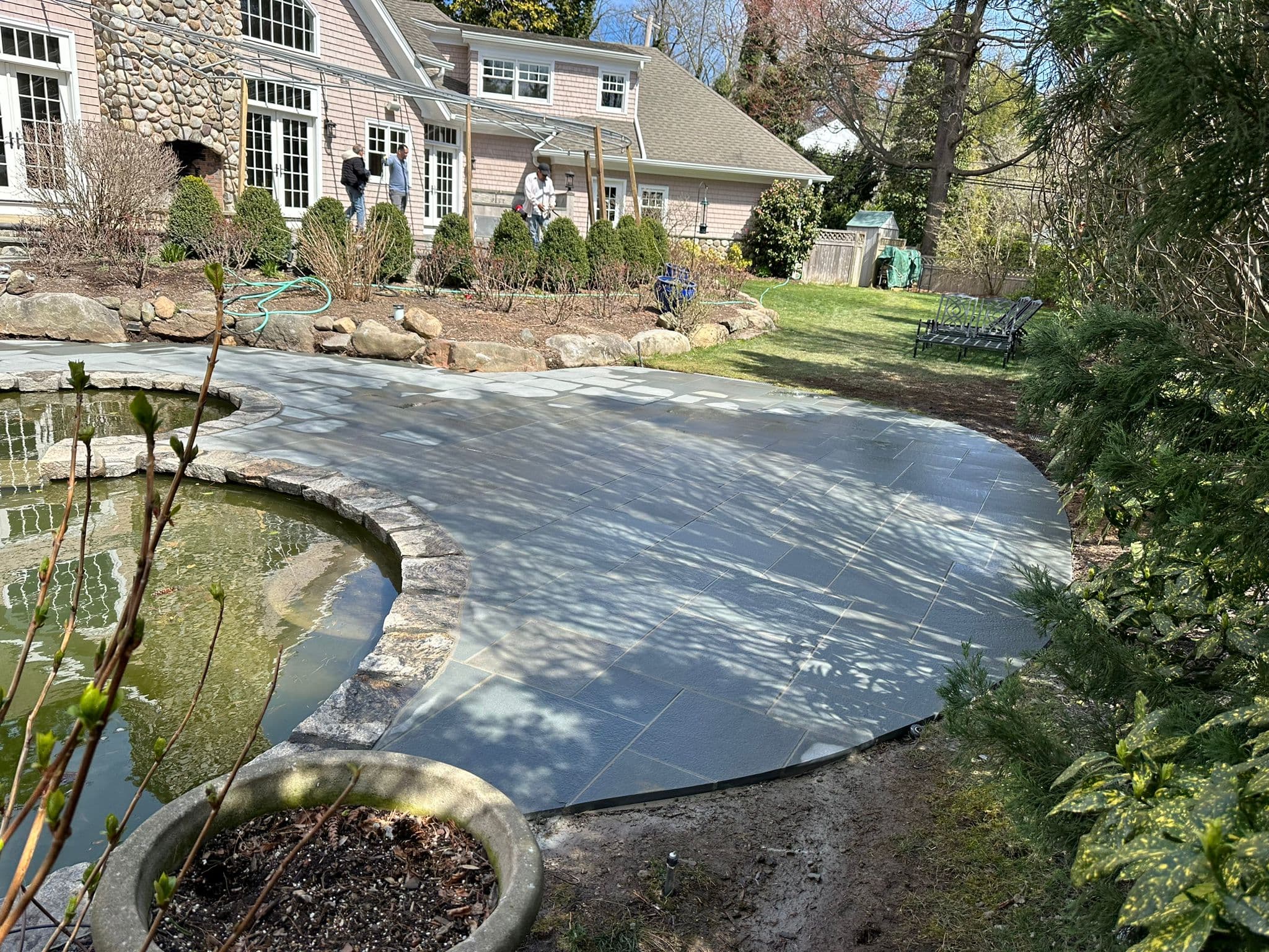 Large grey flagstone patio curving around a garden pond in a landscaped residential backyard.