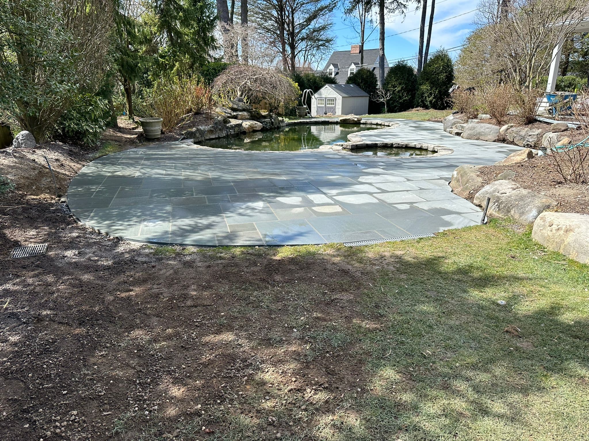 Curved bluestone patio surrounding a garden pond in a landscaped backyard with a shed.